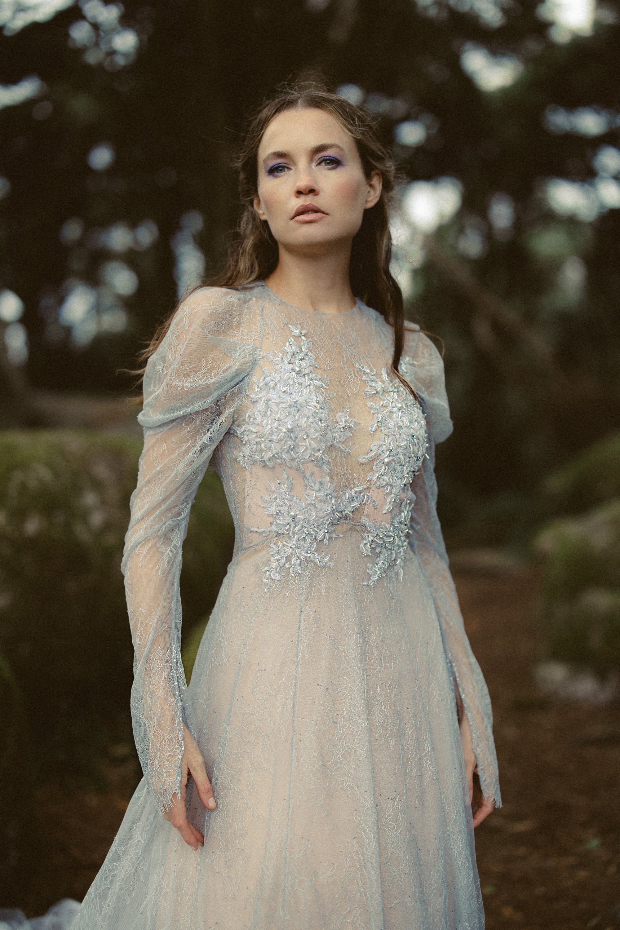 Elystra ethereal wedding dress by MYWONY — portrait of dove-grey floral lace gown with draped long sleeves and beaded embroidery in forest setting