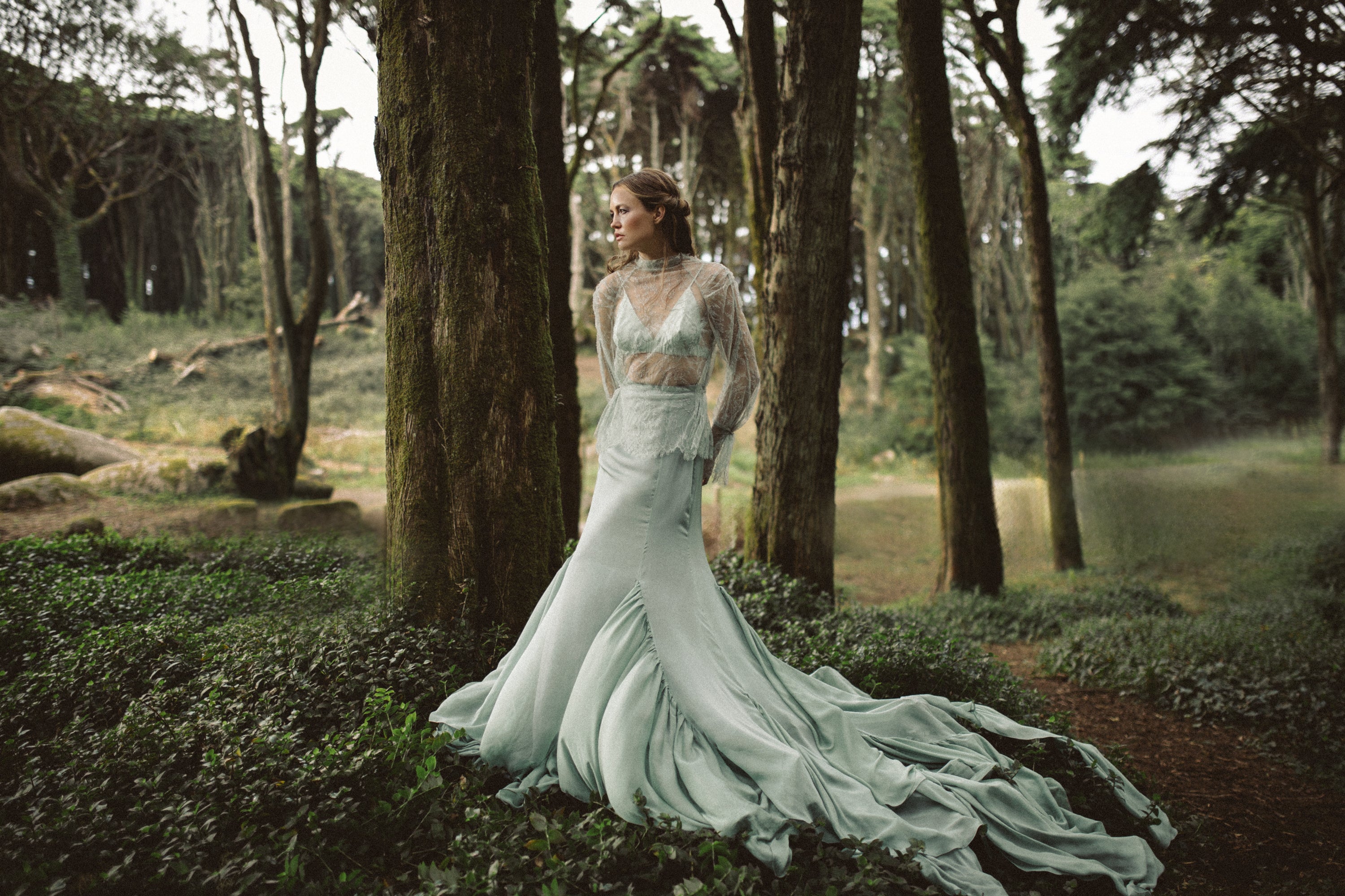 A cinematic wide shot captures the Miryelle modular bridal ensemble in a serene forest setting. The look features a sheer high-neck lace top layered over a pale-green silk triangle bra, paired with a flowing mermaid skirt that cascades into a dramatic chiffon train. The mossy woodland backdrop emphasizes the organic, ethereal aesthetic of this alternative wedding dress.