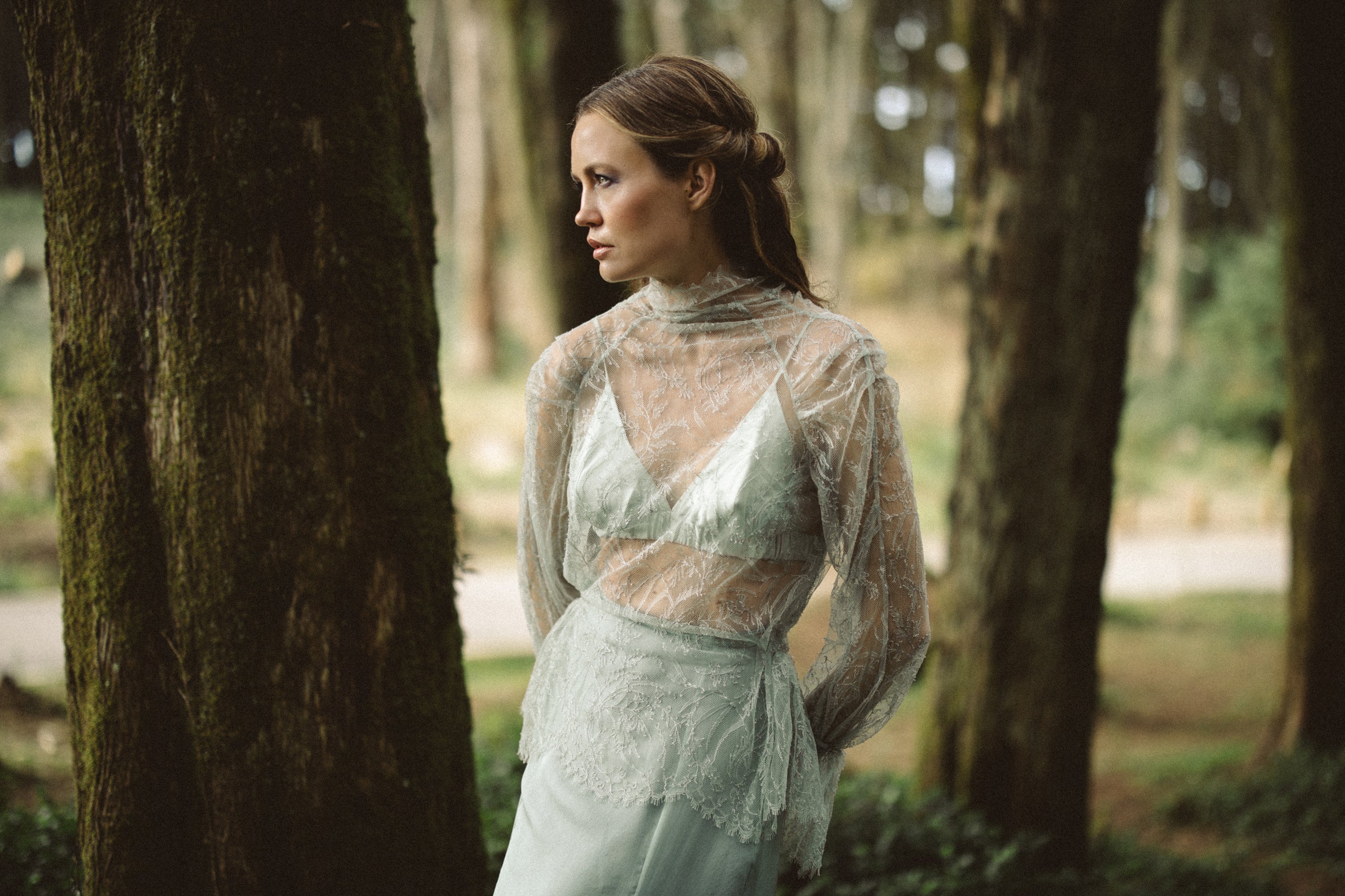 An ethereal portrait showcases the Miryelle lace top, a sheer high-neck blouse featuring delicate floral embroidery and long sleeves. Styled over a pale moss-green silk triangle bra, the ensemble is captured in a moody forest setting, emphasizing the romantic transparency and woodland aesthetic of these modular bridal separates.