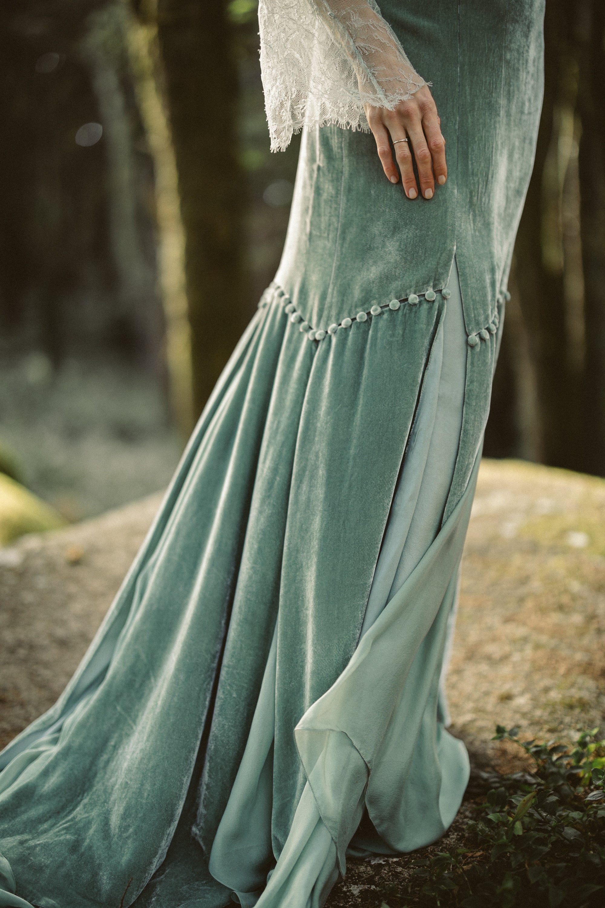 A close-up captures the intricate skirt construction of the Miryelle gown, where pale moss-green silk velour meets layers of ethereal silk chiffon. The image highlights the curved row of covered buttons and the side slit, revealing the fluid movement of the lighter fabrics beneath the structured velvet.