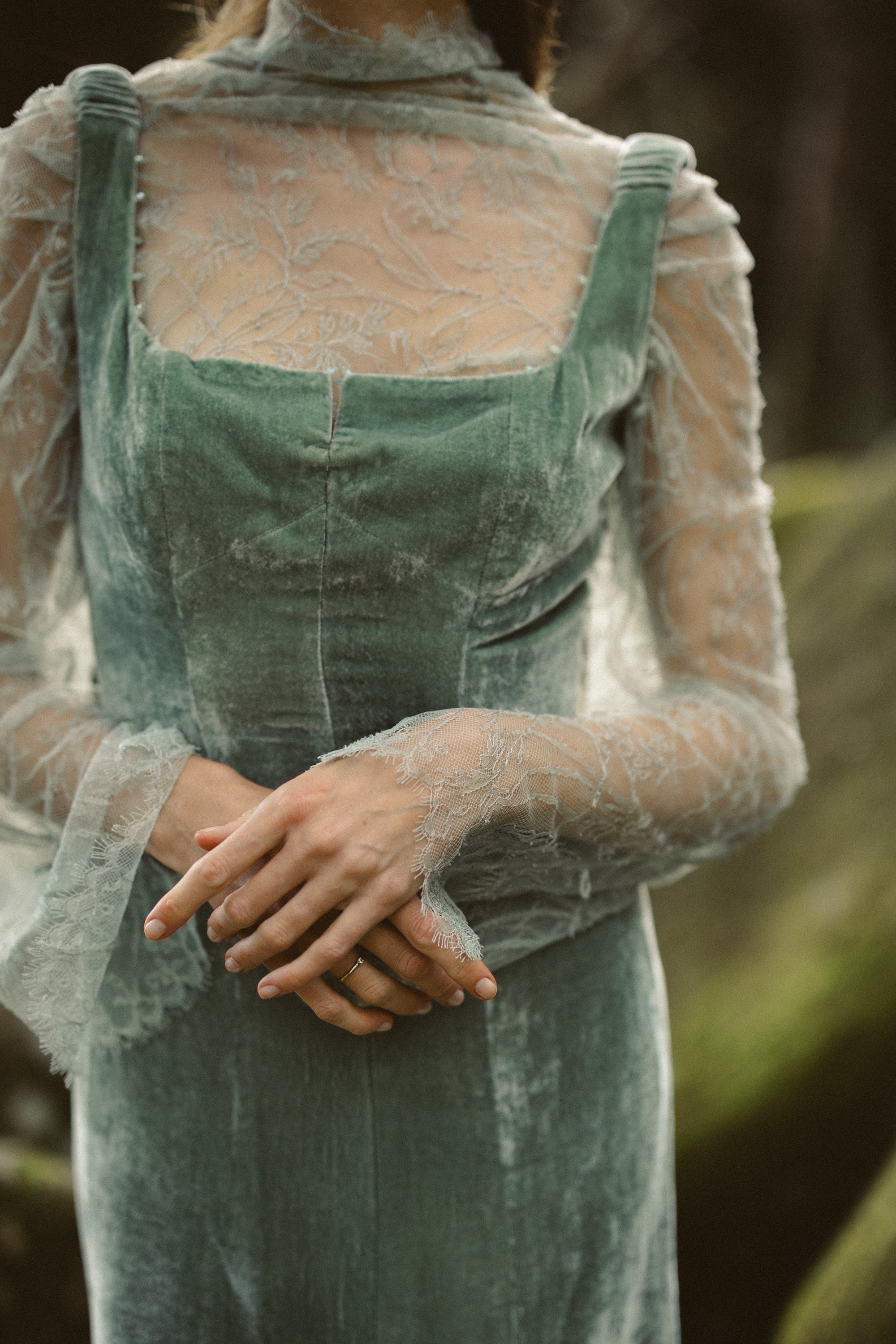 A detailed close-up focuses on the Miryelle bodice, showcasing the plush pale moss-green silk velour against the intricate floral lace underlay. The image highlights the structured square neckline and sheer bell sleeves that drape softly over the model's clasped hands.