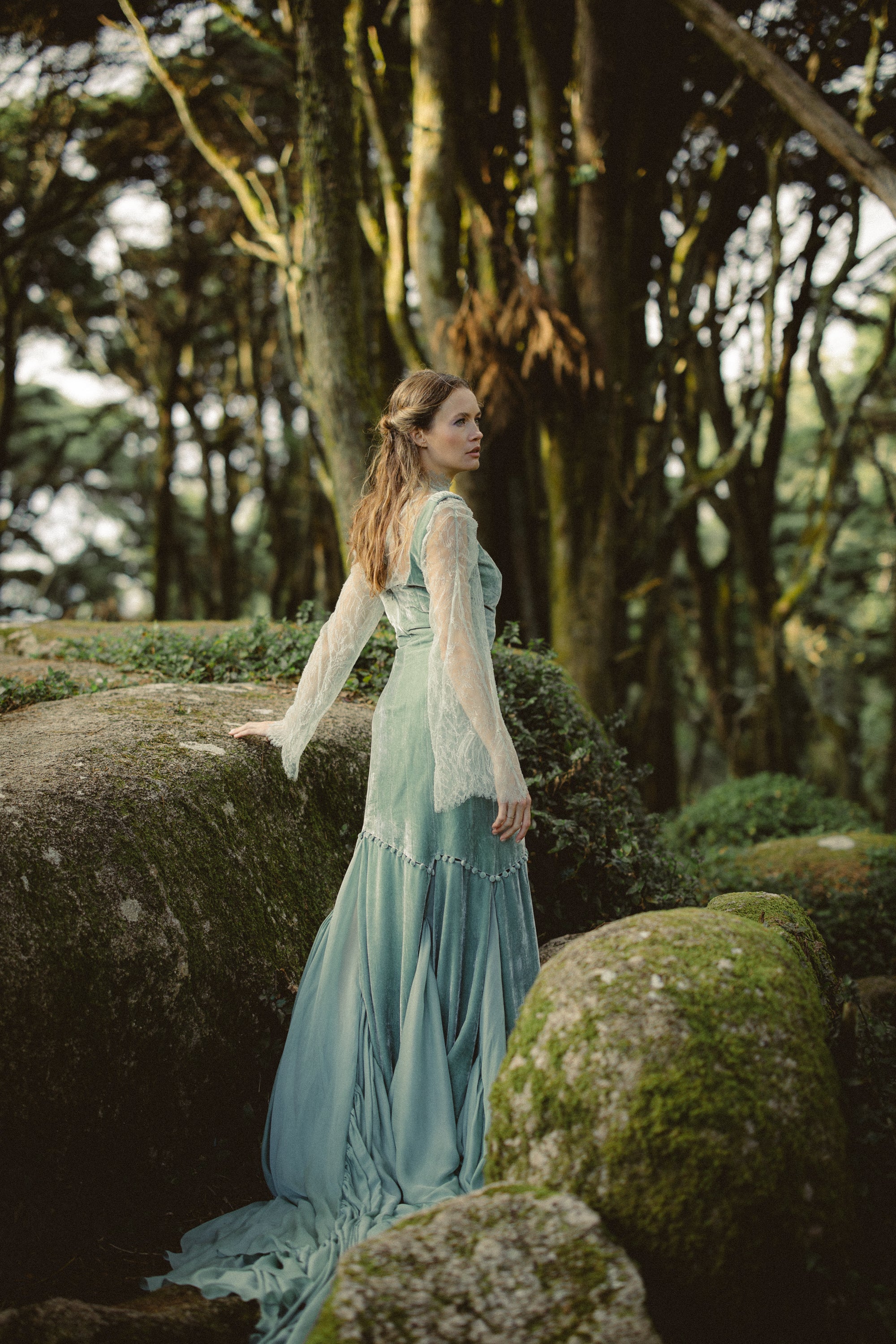A striking full-length rear profile of the Miryelle ensemble set against a backdrop of moss-covered boulders and dense forest trees. The image highlights the textural contrast of the pale moss-green silk velour gown layered over a sheer lace top, featuring dramatic bell sleeves that extend past the wrist. The dress's modular design is showcased at the mid-thigh, where the voluminous mermaid skirt attaches via decorative loops, cascading into a long, fluid train that pools on the forest floor.