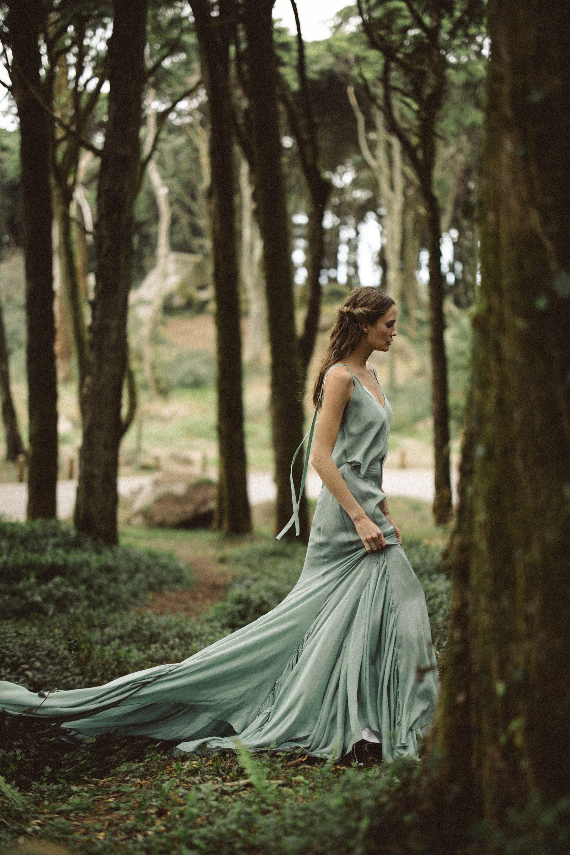 A captivating profile shot captures the fluid motion of the Miryelle silk chiffon ensemble as it glides through the forest. The pale moss-green mermaid skirt creates a sweeping long train, while trailing ribbons highlight the ethereal, bohemian aesthetic of this alternative wedding dress, perfect for brides seeking effortless grace.