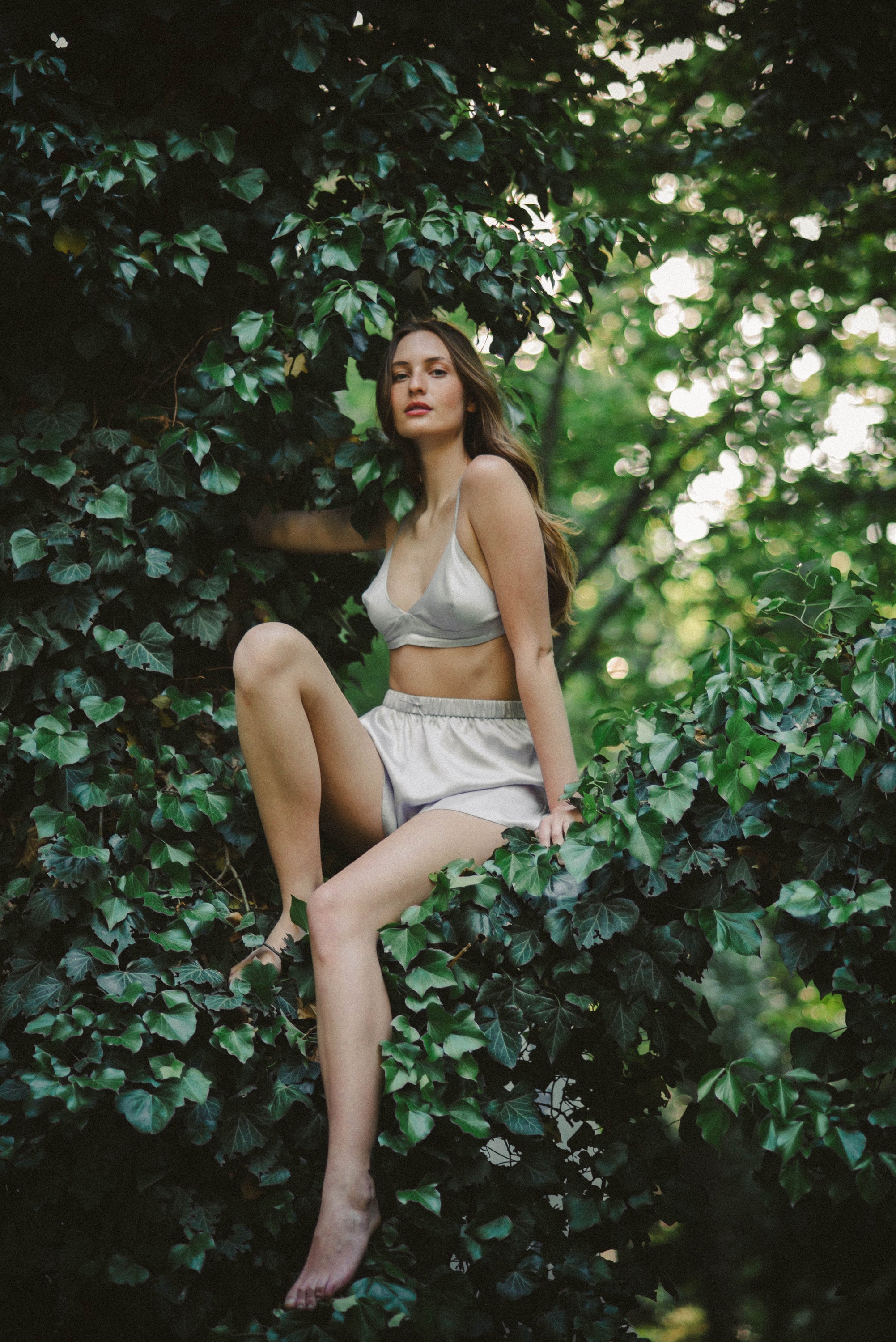 Mýwony mulberry silk bralette and shorts set worn outdoors, model seated among lush garden foliage in editorial campaign shot