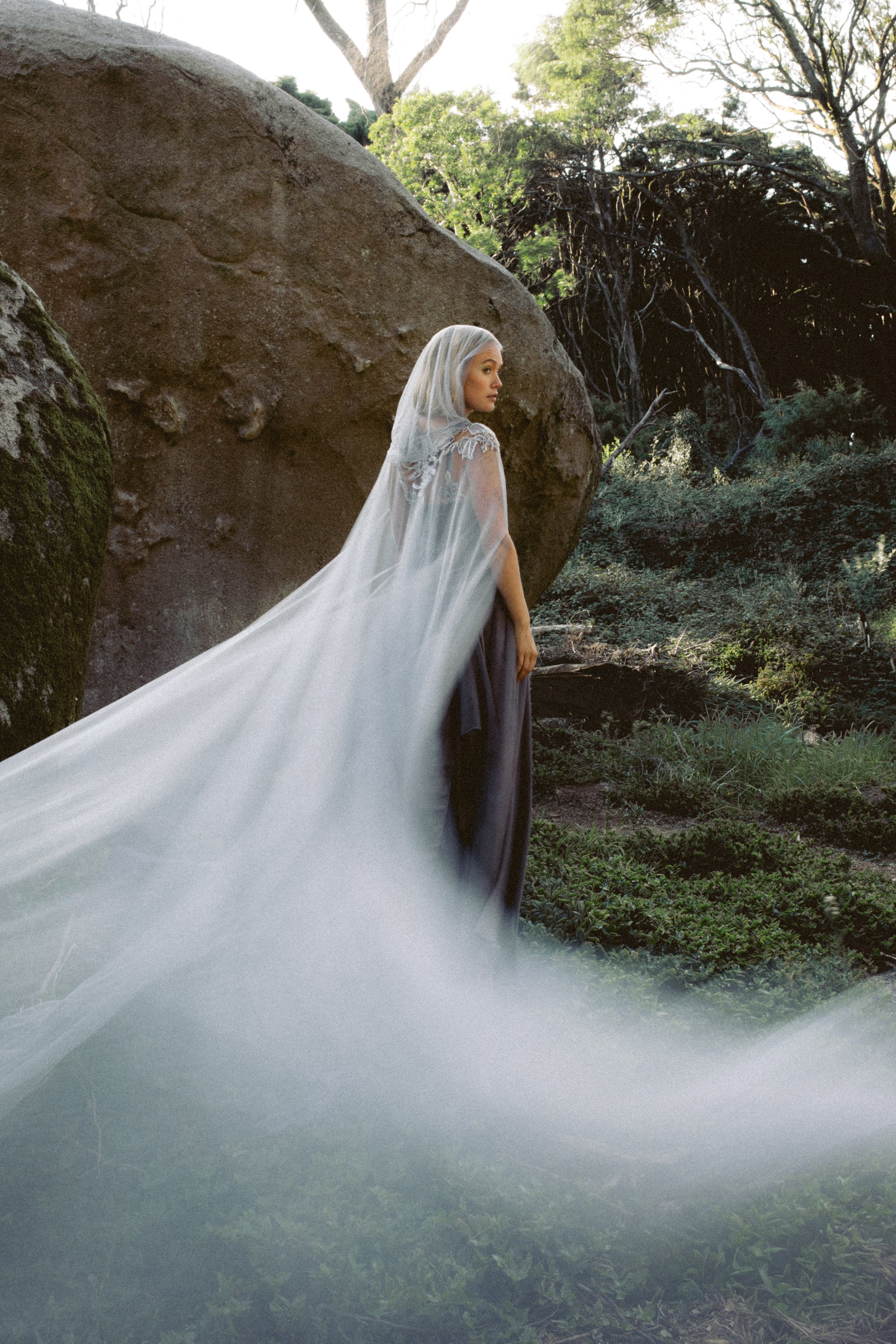 Bride wearing Vea Cape ethereal hooded bridal cape by MYWONY in dove-grey tulle with flowing train, styled in enchanted forest setting for whimsical wedding