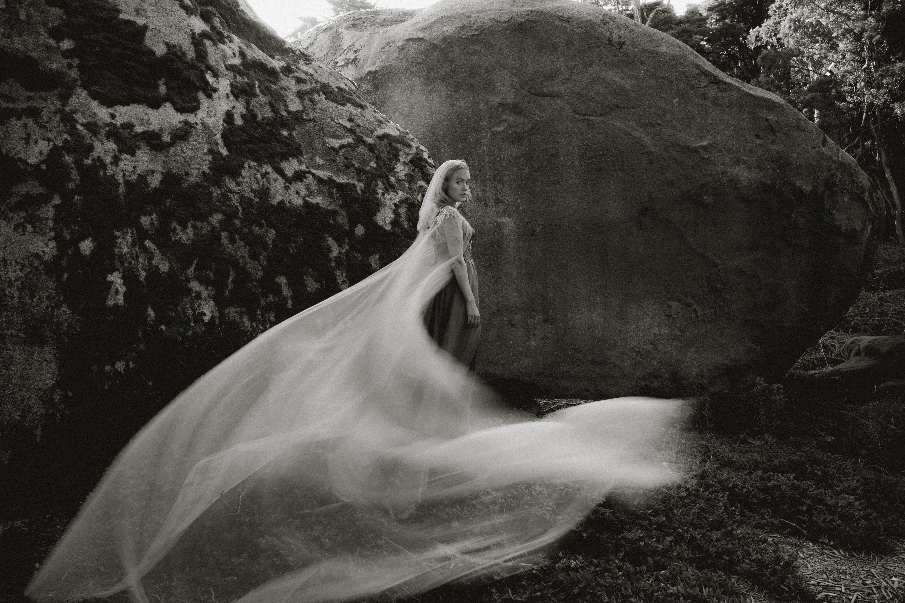 Ethereal black and white photo of Vea Cape custom wedding cape by MYWONY, flowing dove-grey tulle train between forest rocks, whimsical bridal accessory