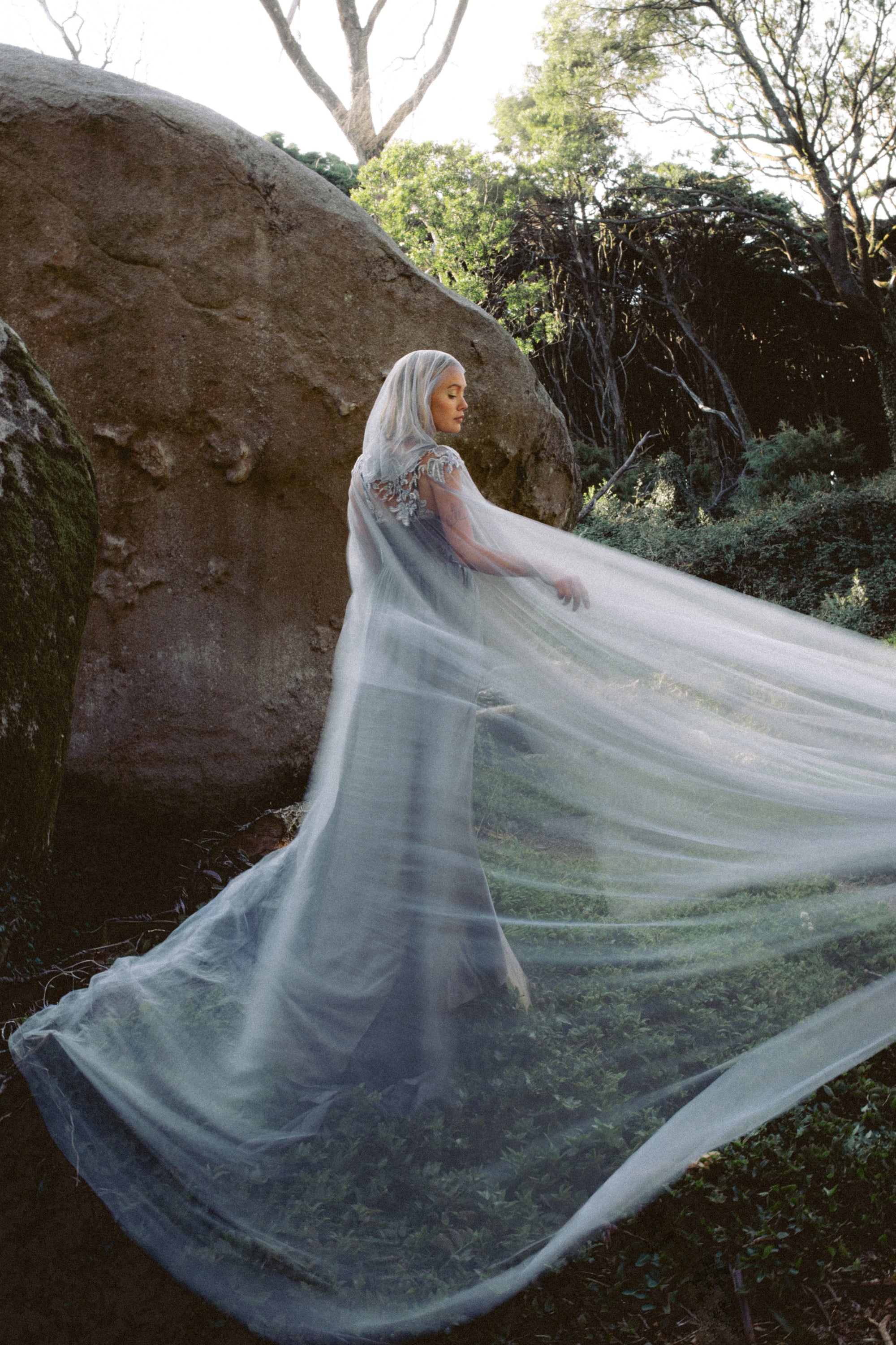 Vea Cape whimsical hooded wedding cape by MYWONY with lavender beaded lace embroidery, bride standing in natural forest setting with flowing tulle train