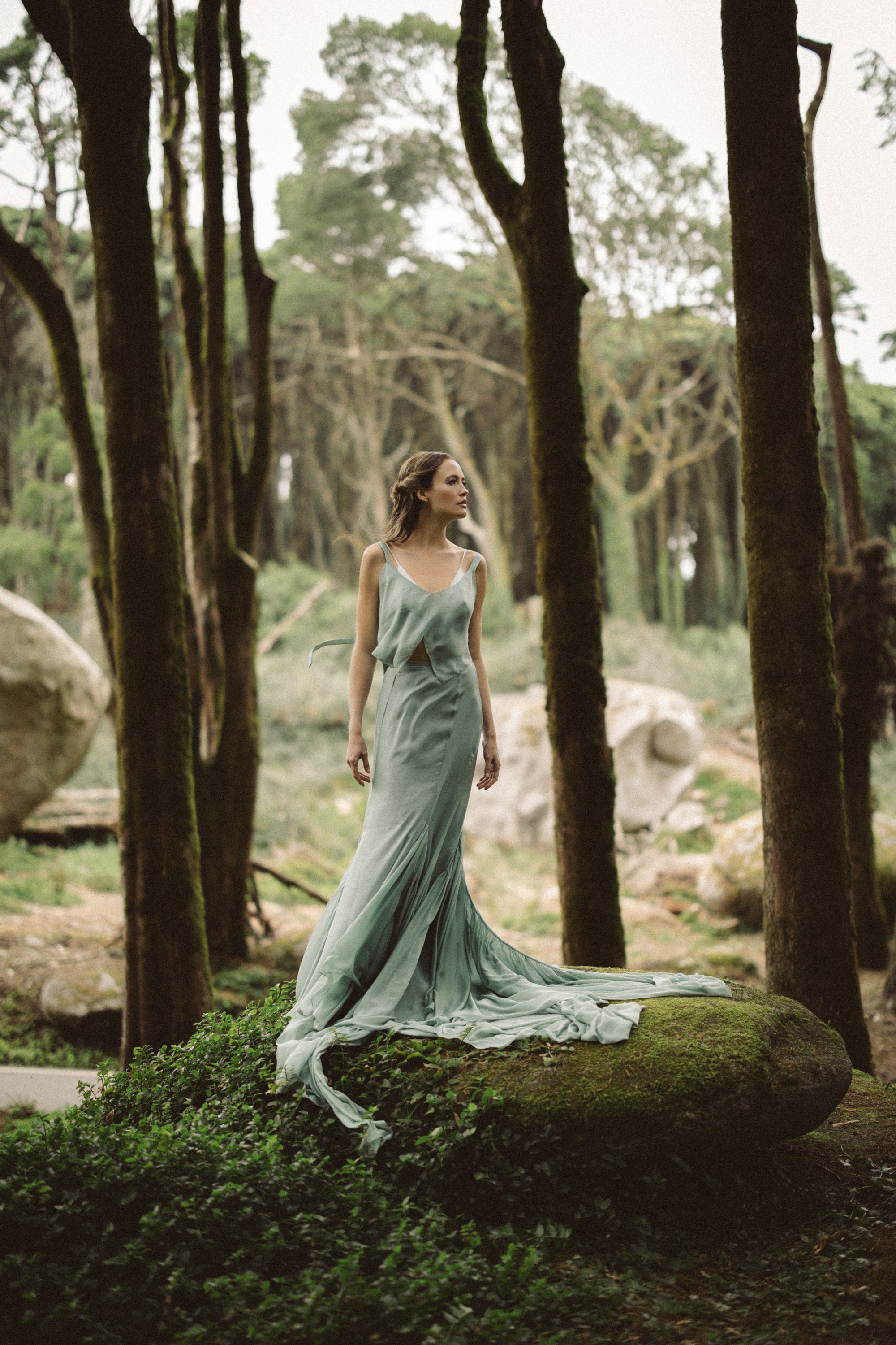 A breathtaking full-length editorial captures the Miryelle modular bridal ensemble in its element, standing atop a moss-covered boulder in an ancient forest. The model showcases the pale moss-green silk chiffon top with its signature geometric four-point hem, paired with a voluminous mermaid skirt that creates a stunning, fluid silhouette. The long chiffon train pools dramatically on the rock, highlighting the ethereal movement of the high-quality silk fabrics. Designed for the non-traditional bride