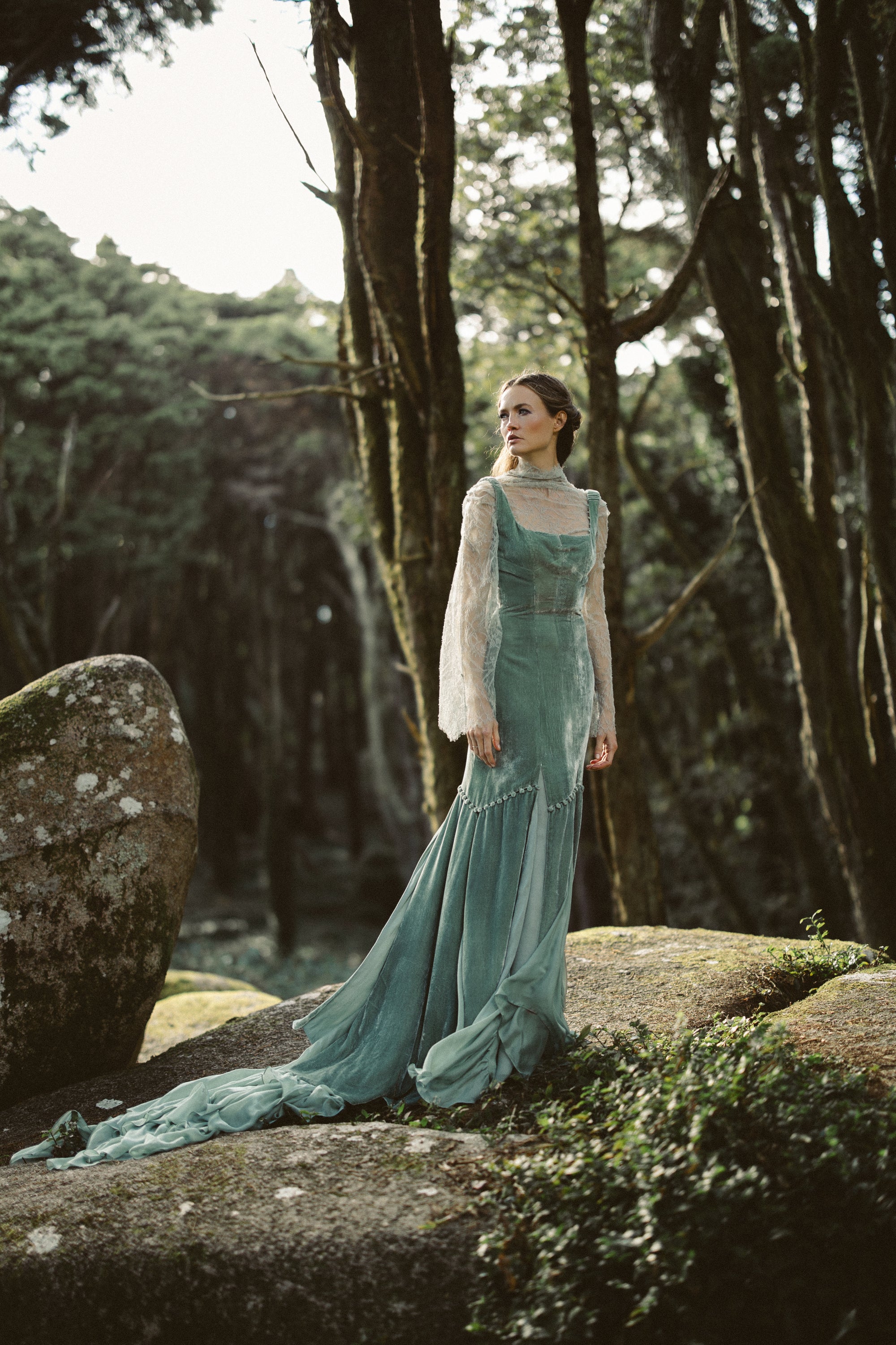 Full-length view of the Miryelle gown in pale moss-green, featuring a silk velour bodice and a voluminous chiffon mermaid skirt cascading over forest rocks.
