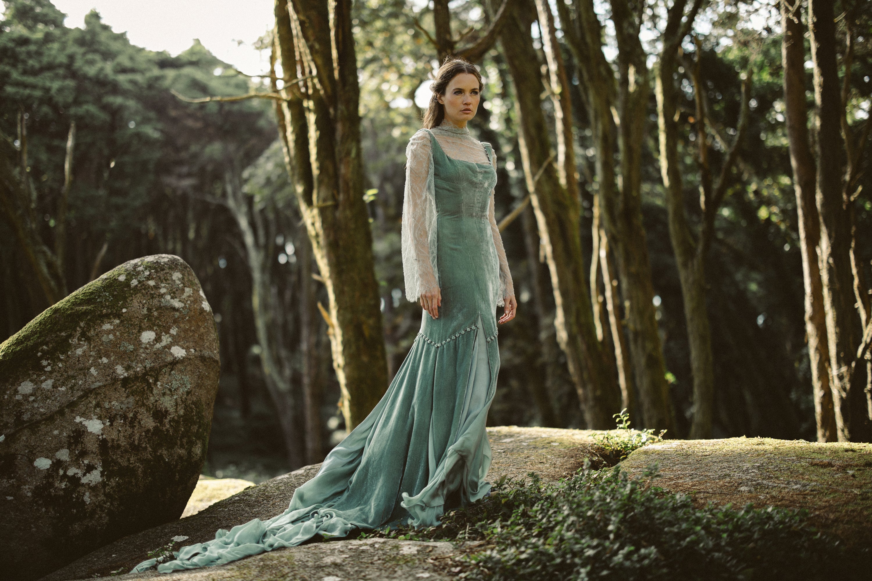 A dramatic full-length portrait showcases the Miryelle ensemble amidst a mossy forest setting. The pale moss-green silk velour gown features a voluminous chiffon train cascading over the rocks, while the underlying sheer lace top with bell sleeves adds delicate texture to the ethereal, nature-inspired silhouette.