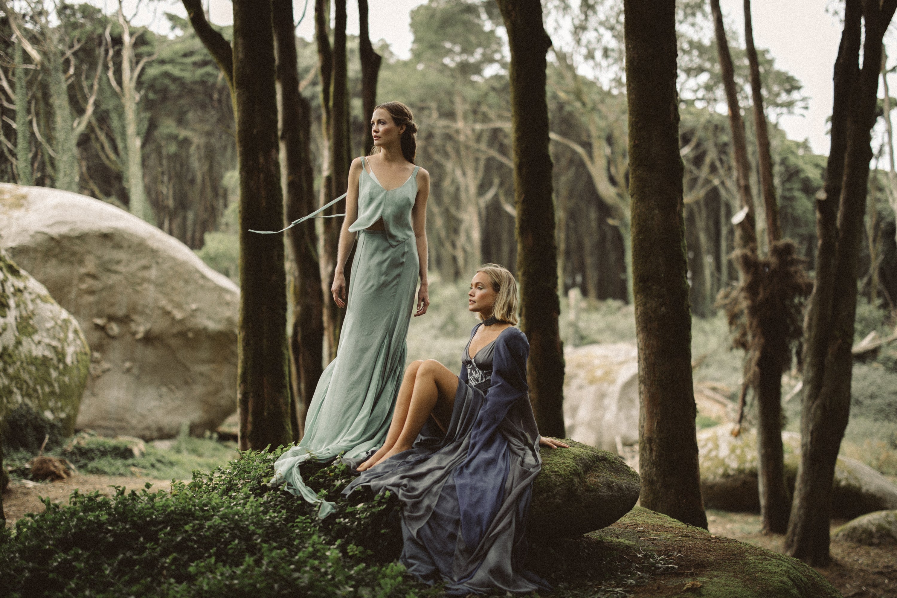 A cinematic forest editorial features two ethereal figures amidst ancient trees. Standing, a model wears the Miryelle ensemble in a relaxed configuration, showcasing the pale moss-green silk chiffon top with flying ribbons and a matching mermaid skirt. Seated on a mossy boulder, the second figure wears a floor-length dark blue-grey gown featuring hand-beaded lavender lace and layered chiffon sleeves, creating a stunning contrast of light and shadow in this woodland fantasy scene.
