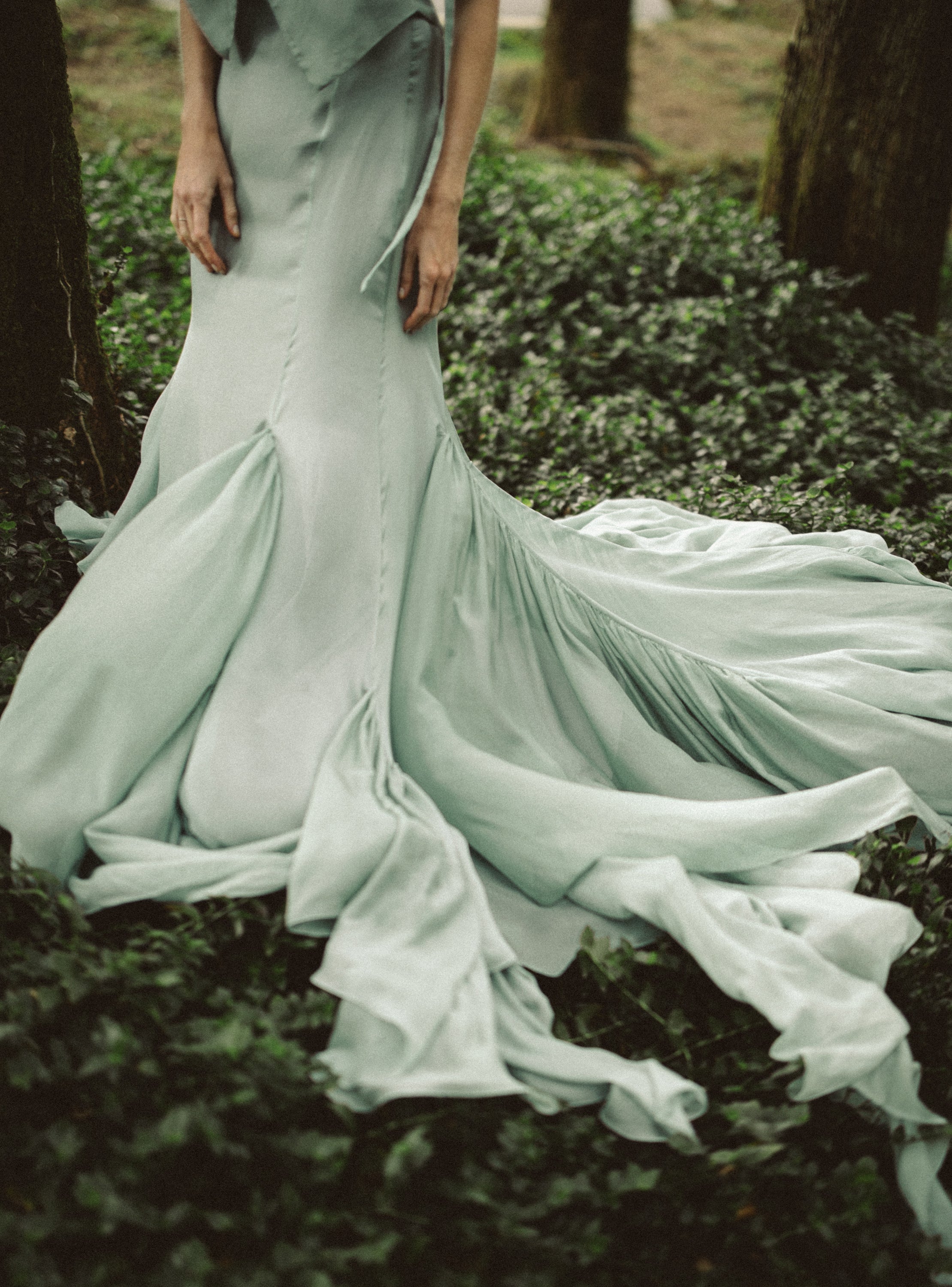 A detail shot emphasizes the dramatic lower silhouette of the Miryelle mermaid skirt, crafted from layers of pale moss-green silk chiffon. The image showcases the skirt's voluminous train cascading over the forest floor, highlighting the fluid movement and intricate paneling of this modular bridal separate.