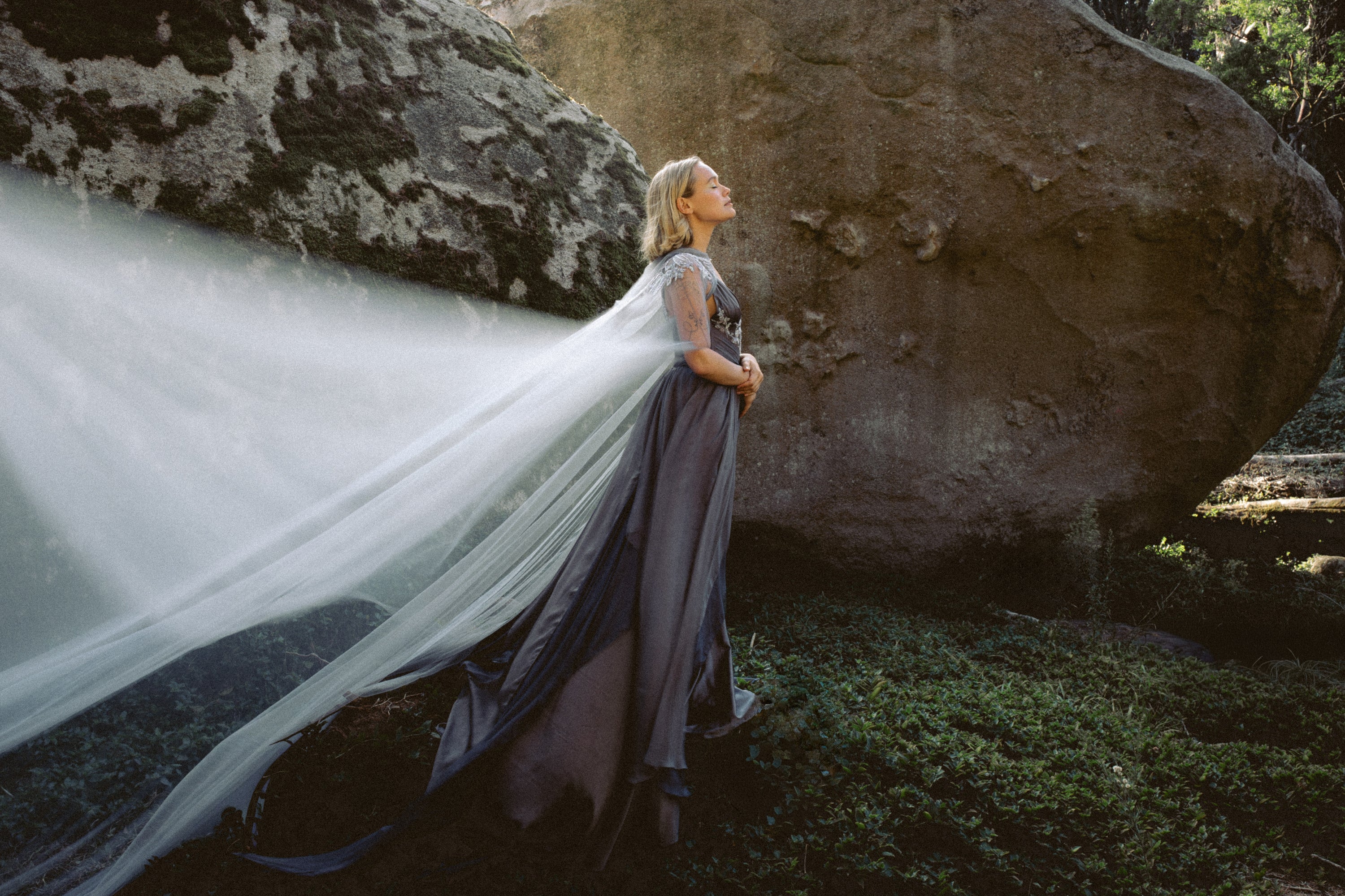 Dramatic side view of Vea Cape custom wedding cape by MYWONY, dove-grey tulle train flowing in forest setting with natural rock backdrop