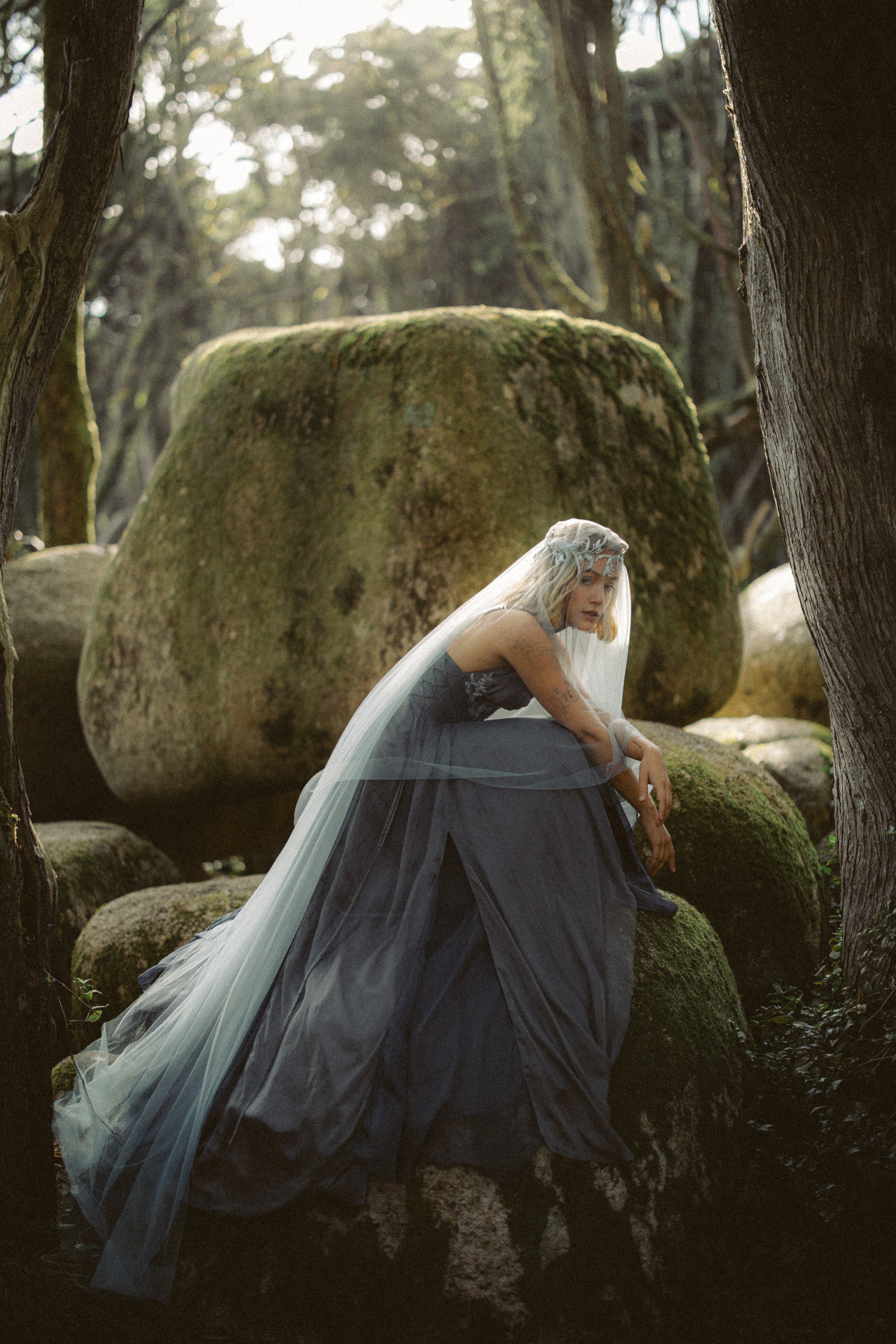 Bride wearing Vea Veil boho wedding veil by MYWONY sitting on moss-covered forest rocks, styled with Vea gown showing ethereal blue tulle layering