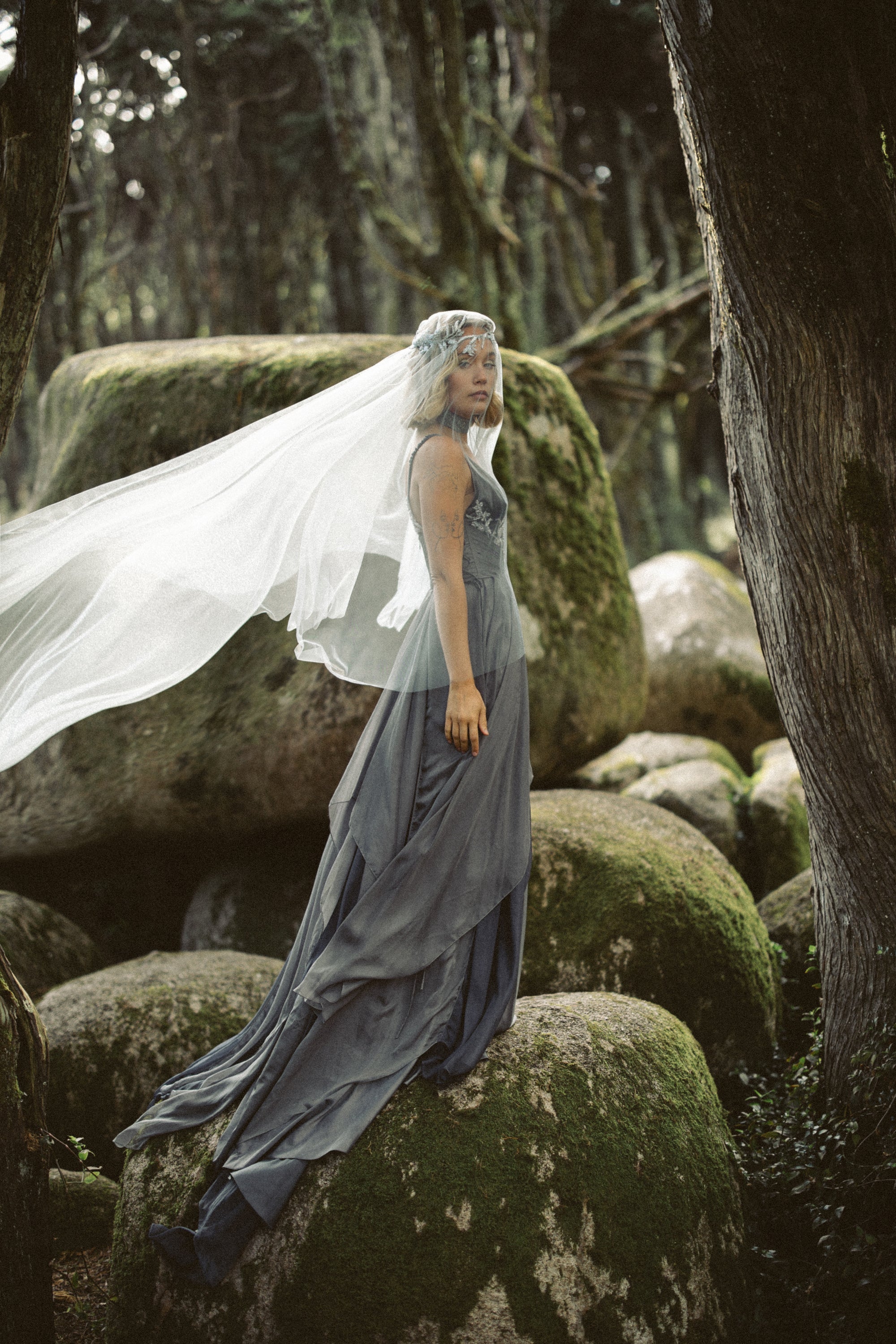 Full-length view of Vea Veil romantic blue tulle wedding veil by MYWONY, bride standing on moss-covered rocks in forest with flowing ethereal drape
